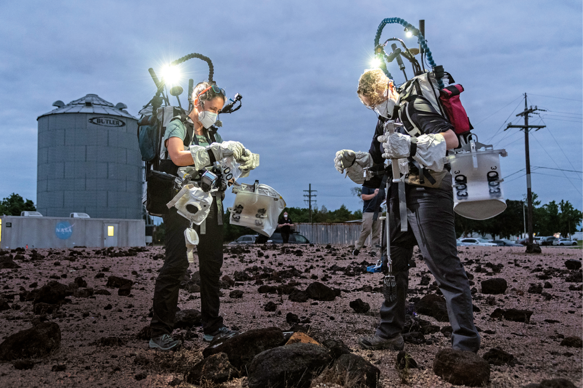 Two people wearing simulated space equipment pick up rocks in NASA test field